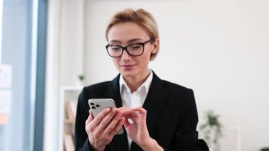 Kendine güvenen yetişkin iş kadını resmi siyah takım elbise giyiyor. Parlak ofis alanında çalışırken akıllı telefon kullanıyor.
