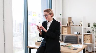 Kendine güvenen yetişkin bir kadın, sarışın, kısa saçlı, resmi takım elbiseli, akıllı telefonla iletişim kuran, modern ofis içi.