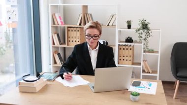 Otuzlarında profesyonel bir kadın modern ofiste masa başında oturmuş belgeleri analiz ediyor ve dizüstü bilgisayarla çalışıyor. Çalışma alanı, üretkenliği ve odaklanmayı artıran doğal ışık, organize raflar içerir..