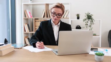 Orta yaşlı, gözlüklü, ofiste dizüstü bilgisayarla kağıt üzerinde çalışan sarışın bir kadın..