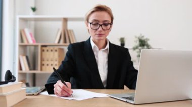 Olgun bir kadın, profesyonel görünümlü, ofis masasında dizüstü bilgisayarla çalışan. Gözlük takıyor, resmi giyiniyor, içeride. İş, teknoloji, konsantrasyon, problem çözme kavramı