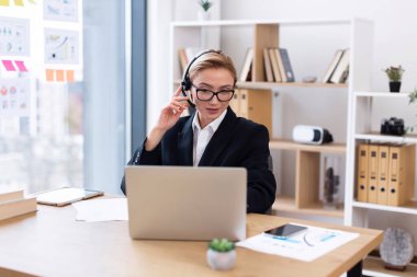 Profesyonel, orta yaşlı, beyaz iş elbiseli kadın modern rezonans ofisinde çalışırken video aracılığıyla iletişim kuruyor. O, masasında bir kulaklık takarken bir laptop kullanıyor..