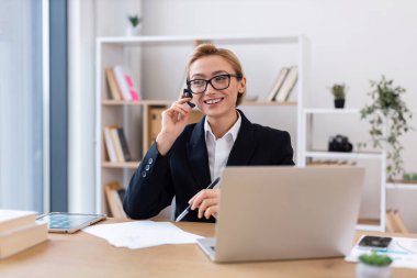 Ofiste çalışan, not yazan ve dizüstü bilgisayar kullanan orta yaşlı bir kadın. Çalışma alanı, üretken ve pozitif bir atmosfer yaratan raflar ve bitkilerle modern ve düzenlidir..