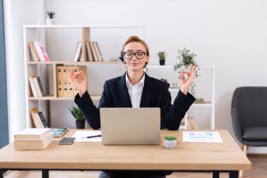 Nilüfer çiçeğiyle ofis masasında meditasyon yapan yetişkin bir kadın, gözlük takıyor ve takım elbise takıyor. Mola süresince iş hayatı dengesini ve farkındalığı destekliyor. Üretimi arttırıyor ve davranışlarını koruyor..