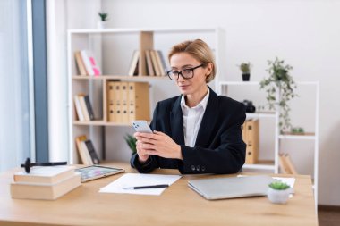 Ofis masasında çalışan gözlüklü bir iş kadını iş aletleriyle çevrili akıllı telefonu kullanıyor. Arkaplanda düzenli raflar olan parlak modern ortam profesyonel atmosfer yaratıyor.