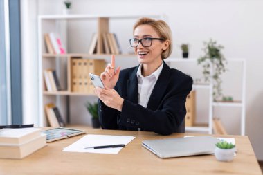 Masada oturan akıllı giyimli iş kadını mutlu bir ifadeyle akıllı telefon kullanıyor. Ofiste profesyonel ortam yaratan organize kitap rafları, kağıtlar ve bitkiler yer alıyor.