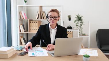 Ofis masasında dizüstü bilgisayarla finansal belgeleri sunan resmi kıyafetli kendine güvenen bir iş kadını. Ofis bitki dekoru arka planda görünür