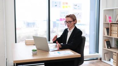 Profesyonel, orta yaşlı, beyaz iş elbiseli kadın modern rezonans ofisinde çalışırken video aracılığıyla iletişim kuruyor. O, masasında bir kulaklık takarken bir laptop kullanıyor..