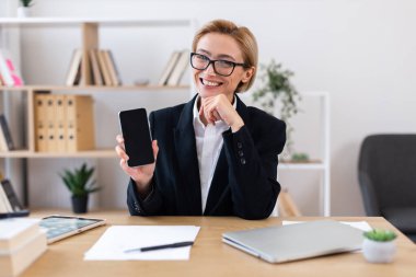 İş kıyafetleri içinde orta yaşlı bir kadın elinde akıllı telefon, teknoloji, takım çalışması, modern ofis gibi kavramlar sunuyor..