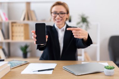Sarı saçlı genç bir kadın ofis ortamında akıllı telefon ekranını sunuyor. Dizüstü bilgisayar, not defteri ve masa başında saksı bitkisi içeriyor. Sahne, profesyonel çalışma ortamında teknoloji kullanımını iletir