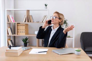 Orta yaşlı, beyaz iş elbiseli bir kadın modern ofis çalışma alanında telefonla konuşuyor.