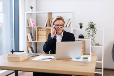 Ofis malzemeleriyle çevrili, resmi takım elbiseli, telefonla konuşan ve laptopta çalışan kendine güvenen yetişkin bir kadın. Verimlilik, çoklu görev ve iş etiğini vurgulayan parlak ofis ortamı.