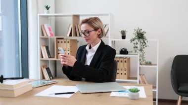 Ofis masasında çalışan gözlüklü bir iş kadını iş aletleriyle çevrili akıllı telefonu kullanıyor. Arkaplanda düzenli raflar olan parlak modern ortam profesyonel atmosfer yaratıyor.