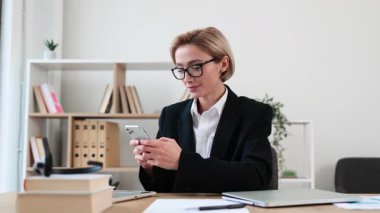 Masada oturan akıllı giyimli iş kadını mutlu bir ifadeyle akıllı telefon kullanıyor. Ofiste profesyonel ortam yaratan organize kitap rafları, kağıtlar ve bitkiler yer alıyor.