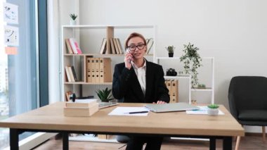 İş yerindeki ofis masasında oturmuş pozitifliğini gösteren telefon konuşmalarından hoşlanan bir kadın. Orta yaşlı kadın, profesyonel kıyafet, gündüz kuşağı, belgeseller, ve çevrede yeşillik.