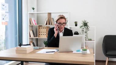 Orta yaşlı, beyaz iş elbiseli bir kadın modern ofis çalışma alanında telefonla konuşuyor.