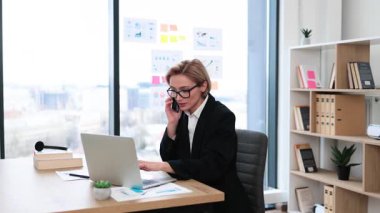 Orta yaşlı bir kadın telefonda konuşuyor, el kaldırarak kutluyor, modern ofiste çalışıyor.