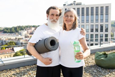 Üst düzey erkek ve kadın çatıda yoga minderi ve su şişesi tutarak aktif yaşam tarzını ve mutluluğunu gösteriyor. Şehir manzarasına karşı sağlıklı yaşam, egzersiz ve bağlantıyı yakalar