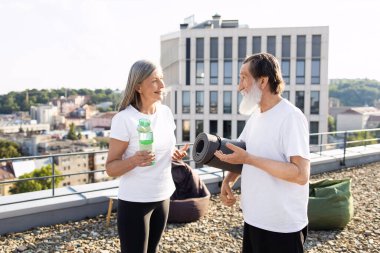 Yaşlı adam ve kadın dışarıda yoga seansına hazırlanıyor. Ellerinde minder ve su şişesi var. Şehrin çatısında gün ışığında gülümsüyorlar..