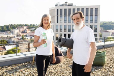 Üst düzey erkek ve kadının açık hava portresi, yoga minderi ve su şişesi, çatı katı arka planı. Yaşlılar için sağlık, spor, açık hava aktiviteleri, aktif yaşam tarzı.