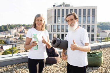 Gülümseyen yaşlı adam ve kadın çatıdaki terasta egzersiz paspası ve su şişesi tutuyor. Aydınlık ve açık bir gün, kentsel geçmiş, spor yapmayı seven yaşlı bireyler için aktif yaşam tarzını teşvik etmek