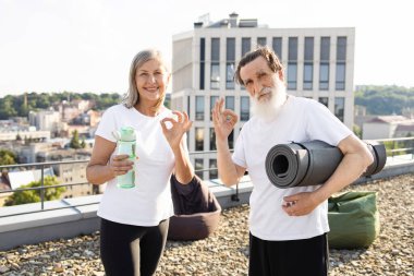 Üst düzey erkek ve kadın çatıda gülümsüyor, yoga ya da fitness aktivitesine hazırlanıyor. Egzersiz gereksinimleri taşırlar ve pozitif enerji sergilerler, sağlıklı yaşam tarzını teşvik ederler ve aktif kalırlar.
