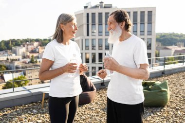 Üst düzey beyaz erkek ve kadın aktif spor giyim kuşamında açık çatıda şişe su içiyorlar. Görüntü aktif yaşam tarzı, açık hava fitness, egzersiz sonrası ferahlatıcı, şehir arkaplanını gösteren.
