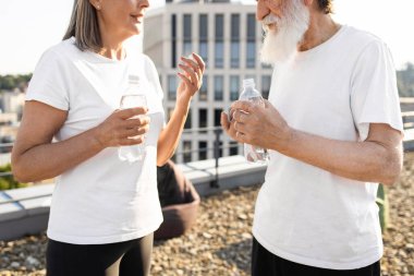 Dışarıda iki kıdemli kişi günlük kıyafetleriyle çatıda şişelenmiş suyun keyfini çıkarıyor. Susuzluk, rahatlama, sağlıklı yaşam tarzı. Yaşlılar, cinsiyetler, açık hava.
