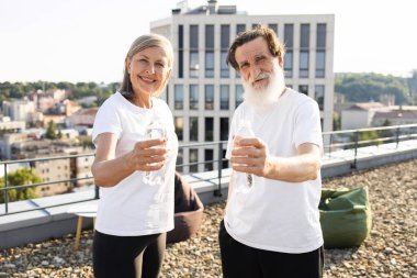 Sakallı yaşlı bir adam ve egzersizden sonra çatıda elinde şişe suyla gülümseyen bir kadın. Kentsel altyapı, gündüz ayarı, ikilisi emeklilikte sağlık, su ve canlılığı simgeliyor.