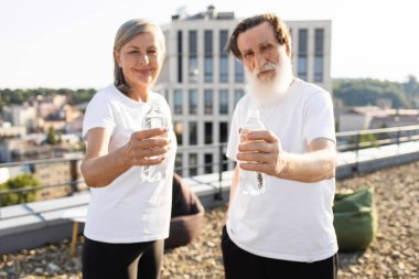 Yaşlı adam ve kadın ellerinde şişe suyla şehrin çatısında temiz havanın tadını çıkarırken idmandan sonra spor sonrası açık hava aktivitelerini temsil eden beyaz gömlekler giyiyorlar.