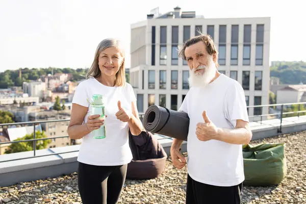 Gülümseyen yaşlı adam ve kadın çatıdaki terasta egzersiz paspası ve su şişesi tutuyor. Aydınlık ve açık bir gün, kentsel geçmiş, spor yapmayı seven yaşlı bireyler için aktif yaşam tarzını teşvik etmek