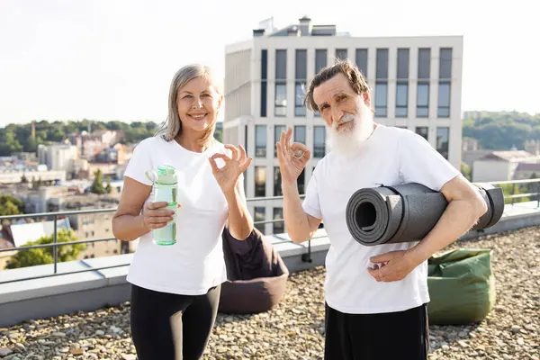 Üst düzey erkek ve kadın çatıda gülümsüyor, yoga ya da fitness aktivitesine hazırlanıyor. Egzersiz gereksinimleri taşırlar ve pozitif enerji sergilerler, sağlıklı yaşam tarzını teşvik ederler ve aktif kalırlar.