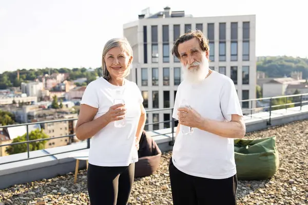 Kıdemli adam ve kadın antrenmandan sonra çatıda durup su içiyorlar. Şehir geçmişi, sağlıklı bir yaşam tarzı var. Sahne günlük spor kıyafetleri, güneşli bir gün, çatı katı, egzersiz sonrası...