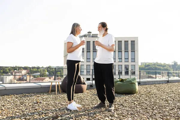 Üst düzey bir adam ve kadın şehrin çatısında dikiliyor şişe su içiyor, açık havada güneş ışığı altında günlük giysiler giyiyor.