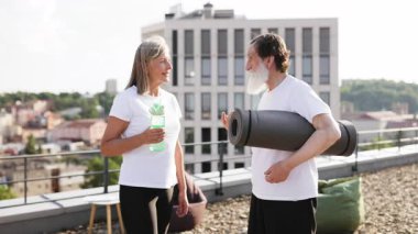 Yaşlı adam ve kadın dışarıda yoga seansına hazırlanıyor. Ellerinde minder ve su şişesi var. Şehrin çatısında gün ışığında gülümsüyorlar..
