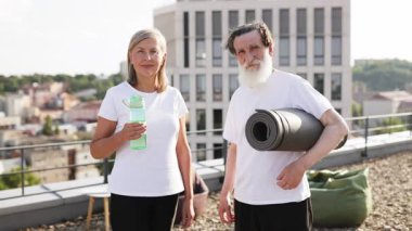 Üst düzey erkek ve kadın çatıda gülümsüyor, yoga ya da fitness aktivitesine hazırlanıyor. Egzersiz gereksinimleri taşırlar ve pozitif enerji sergilerler, sağlıklı yaşam tarzını teşvik ederler ve aktif kalırlar.