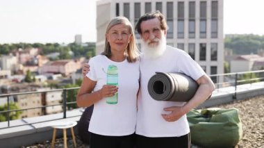 Üst düzey erkek ve kadın çatıda yoga minderi ve su şişesi tutarak aktif yaşam tarzını ve mutluluğunu gösteriyor. Şehir manzarasına karşı sağlıklı yaşam, egzersiz ve bağlantıyı yakalar