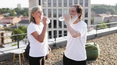 Kıdemli adam ve kadın antrenmandan sonra çatıda durup su içiyorlar. Şehir geçmişi, sağlıklı bir yaşam tarzı var. Sahne günlük spor kıyafetleri, güneşli bir gün, çatı katı, egzersiz sonrası...
