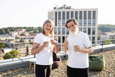 Yaşlı bir adam ve kadın güneşli bir günde ellerinde su şişeleriyle dışarıda dikiliyorlar. Çatı katında gündelik kıyafet. Egzersiz sonrası mutlu, sağlıklı görünüyorlar..