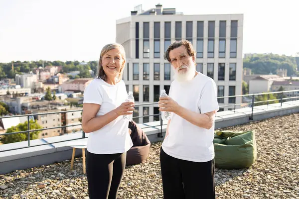 Güneşli bir sabahta şehir çatısı egzersizinden sonra susuz kalan yaşlı bir çift. Günlük spor kıyafetleri giyiyorlar, gülümsüyorlar ve refahın tadını çıkarıyorlar. Şehir manzarası ve mavi gökyüzü kentsel atmosfer sağlar.