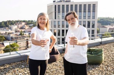 Çatıda elinde su şişeleri olan yaşlı bir adam ve kadın egzersizden sonra gülümsüyor. Açık hava şehir manzarası sağlık ve birlikteliği vurgulayan fitness ve hidrasyon kavramını gösteriyor