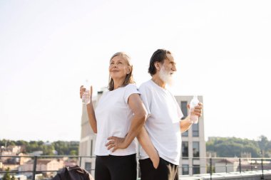 Kıdemli çift antrenmandan sonra çatıda su içiyor. Günlük kıyafetlerle, güneşli bir günde. Sağlıklı yaşam tarzını, sağlığını ve su ihtiyacını arttırır..