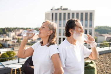 Beyazlar içinde şişelenmiş su içen yaşlı bir adam ve kadın. Kentsel arka plana sahip çatı ayarları fiziksel aktivite ve sağlıklı kalma anlamına gelir.
