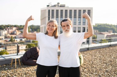 Yaşlı erkek ve kadın çatıda yan yana duruyor, kollarını esnetiyor, aktif yaşam tarzını ve sağlık bilincini destekliyor, olumlu duygular, neşe ve fiziksel kondisyon gösteriyor..