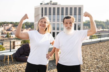 Beyaz sakallı yaşlı bir adam ve beyaz gömlekli bir kadın dışarıda pazılarını gösteriyor.