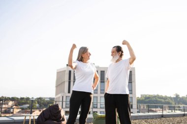 Yaşlı adam ve kadın çatıda kollarını esnetiyor, aktif sağlıklı yaşam tarzını gösteriyor, kendine güveni ve gücü simgeliyor.