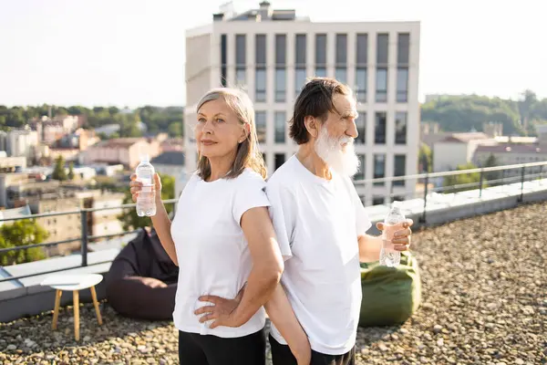 İki kıdemli birey, erkek ve kadın, eğitimden sonra çatıda su şişesi tutuyorlar. Günlük spor kıyafetleri, arka planda şehir manzarası. Aktif sağlıklı yaşam tarzı ve susuz kalma kavramı.