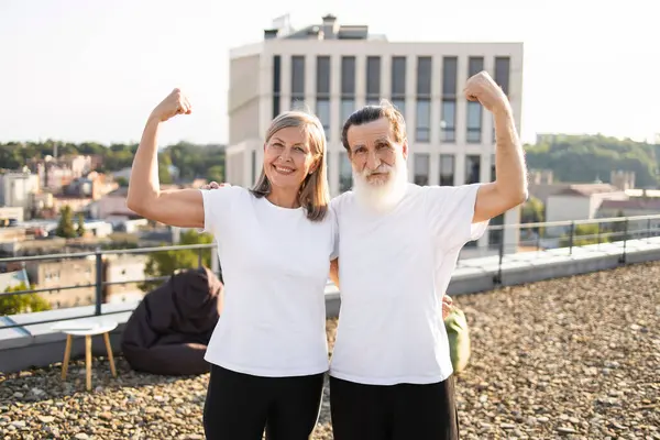 Yaşlı erkek ve kadın çatıda yan yana duruyor, kollarını esnetiyor, aktif yaşam tarzını ve sağlık bilincini destekliyor, olumlu duygular, neşe ve fiziksel kondisyon gösteriyor..