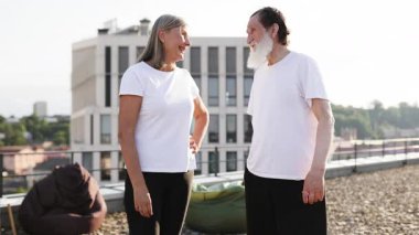 Yaşlı erkek ve kadın çatıda yan yana duruyor, kollarını esnetiyor, aktif yaşam tarzını ve sağlık bilincini destekliyor, olumlu duygular, neşe ve fiziksel kondisyon gösteriyor..