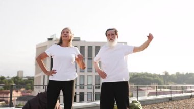 Yaşlı adam ve kadın, açık havada, çatıda, parlak güneş ışığında esneme egzersizleri yapıyorlar. Sağlık, hareketlilik, günlük aktivite, mutlu yaşam tarzı vurgulanıyor.